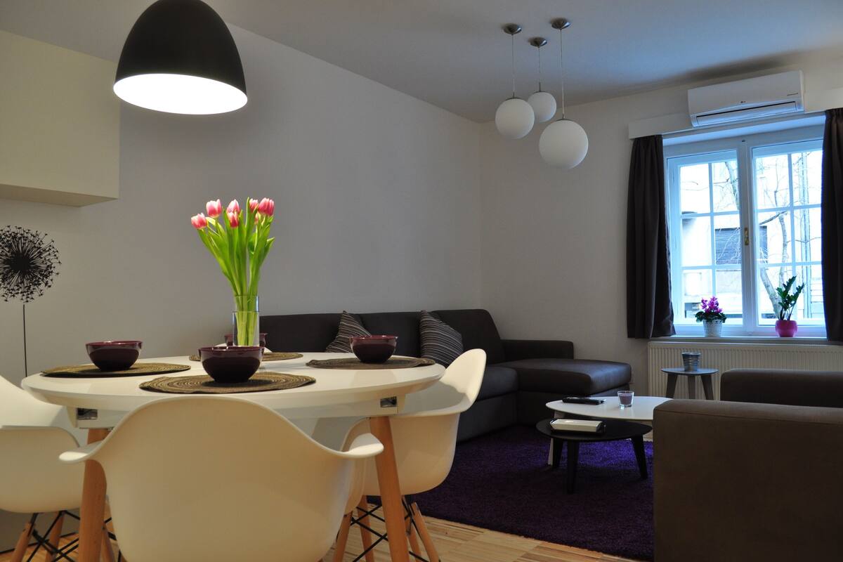 Un salon moderne avec une table à manger ronde, des chaises blanches, un canapé noir et une table basse. Un vase rempli de fleurs roses est posé sur la table à manger.