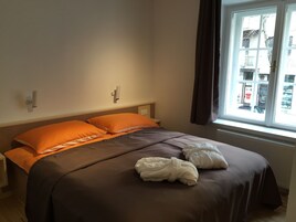 Een net opgemaakt bed met oranje en bruine beddengoed, een raam met uitzicht op gebouwen, en een wandlamp.