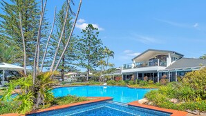 A swimming pool with a metal ladder, surrounded by tropical plants and a modern house with a balcony. - Luxury condo, ocean views, sea breeze, pool, tennis court, minutes to cafes (Sapphire Beach)
