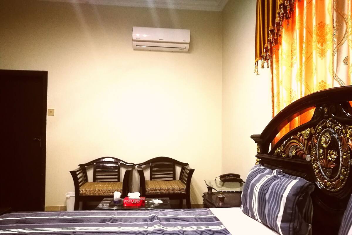 Habitación de hotel con cama, dos sillas, una mesita con una caja de pañuelos y un aire acondicionado en la pared.