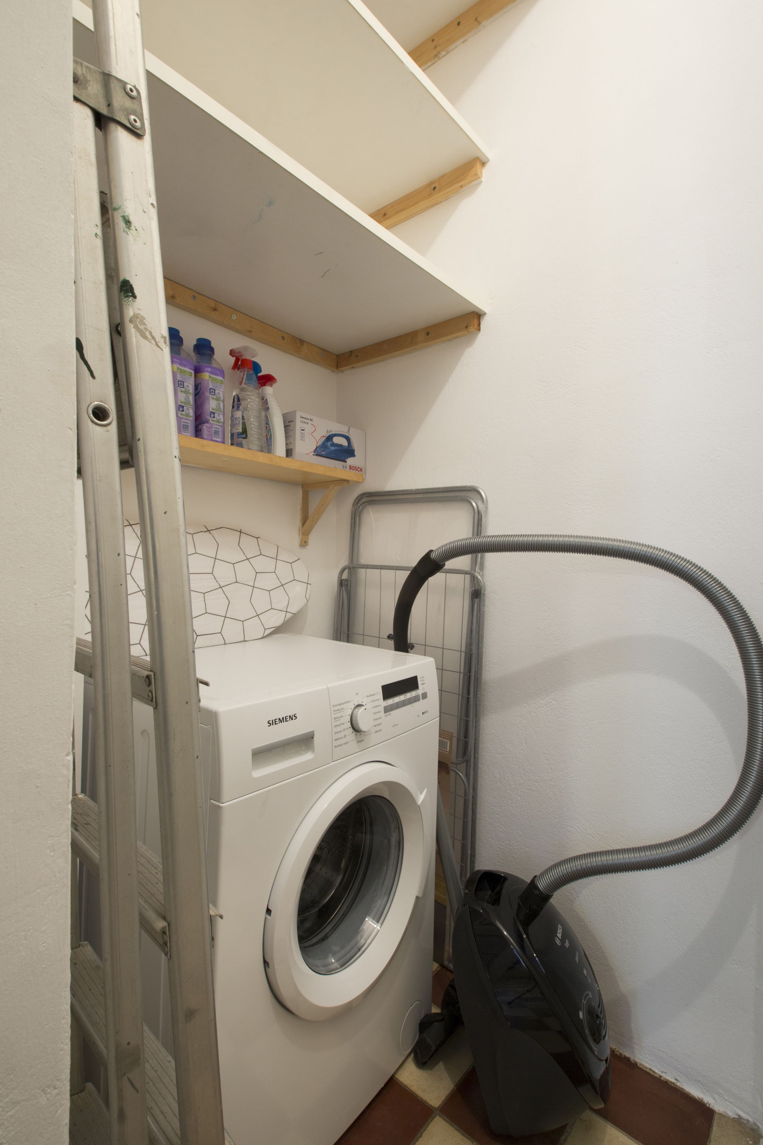 A compact laundry room with a front-loading washing machine, a vacuum cleaner, and a shelf with cleaning supplies.
