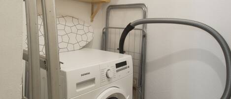 A compact laundry room with a front-loading washing machine, a vacuum cleaner, and a shelf with cleaning supplies.