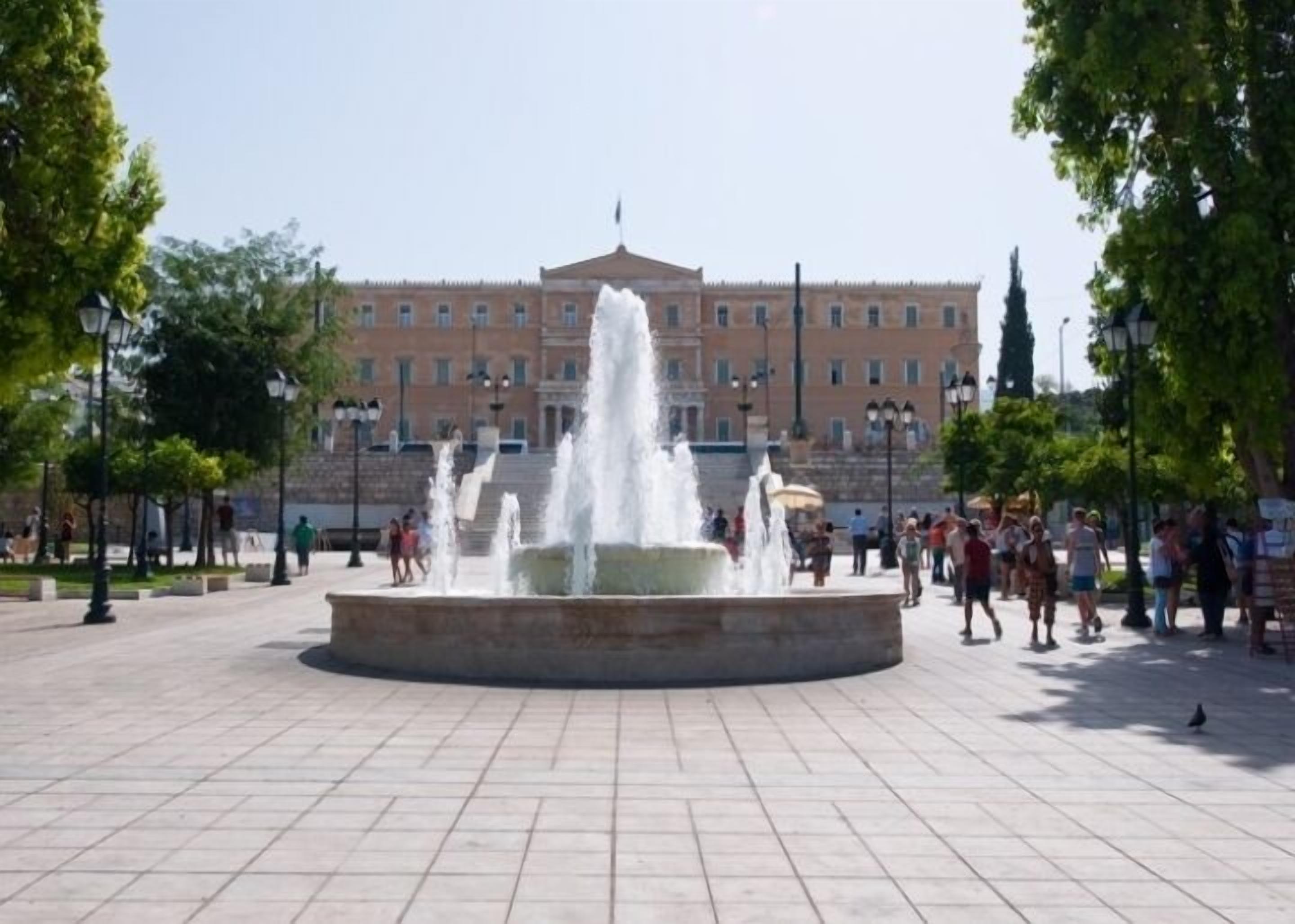 Une place publique avec une fontaine centrale, entourée d’arbres et dominée par un grand bâtiment en arrière-plan.