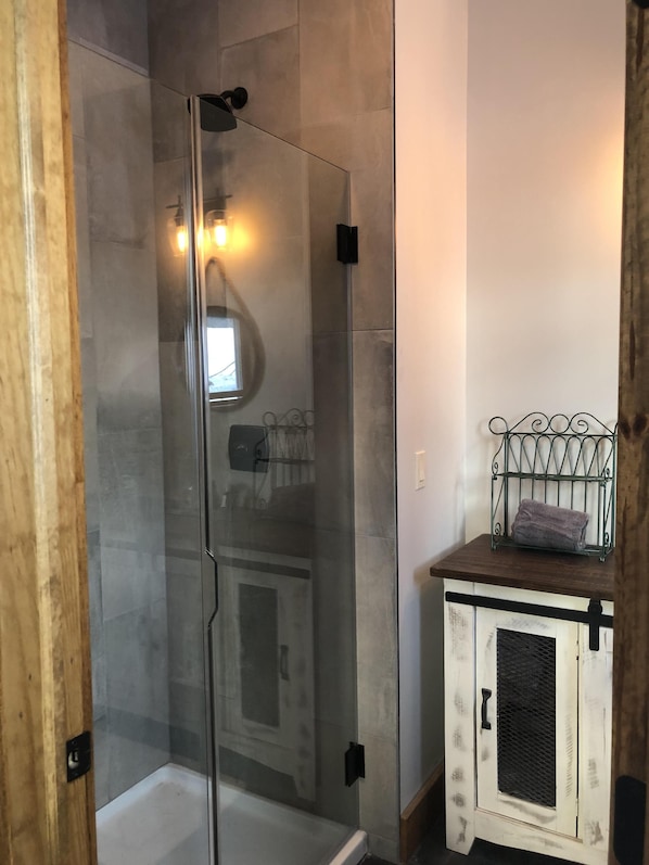 A glass-enclosed shower with a white door, a small wooden cabinet with a metal shelf, and a wall-mounted light fixture. - "New Home" Idyllic Mt Views ,Pond & Horse farm views 2 min to Town (Windham)