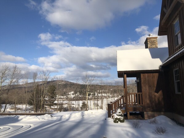 A wooden cabin with a snow-covered roof, a wooden deck, and a view of a snowy landscape with bare trees and distant hills. - "New Home" Idyllic Mt Views ,Pond & Horse farm views 2 min to Town (Windham)