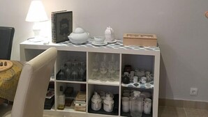 A dining area with a white table, a chair, a lamp, and a shelf with glassware and teapots. - Casa Rural Las Violetas (Villanueva de los Infantes)