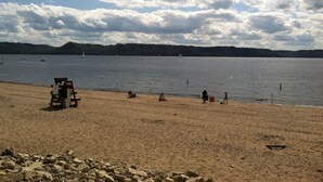 A beach with a chair and a table, a sandy shore, and a calm lake with boats in the distance. - Stunning view of Lake Pepin! 1/2 mile to Villa Bellezza (Pepin)