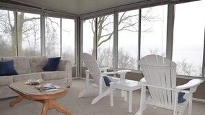 A cozy living room with a beige sofa, a wooden coffee table, and white Adirondack chairs. Large windows offer a view of trees and a cloudy sky. - Stunning view of Lake Pepin! 1/2 mile to Villa Bellezza (Pepin)