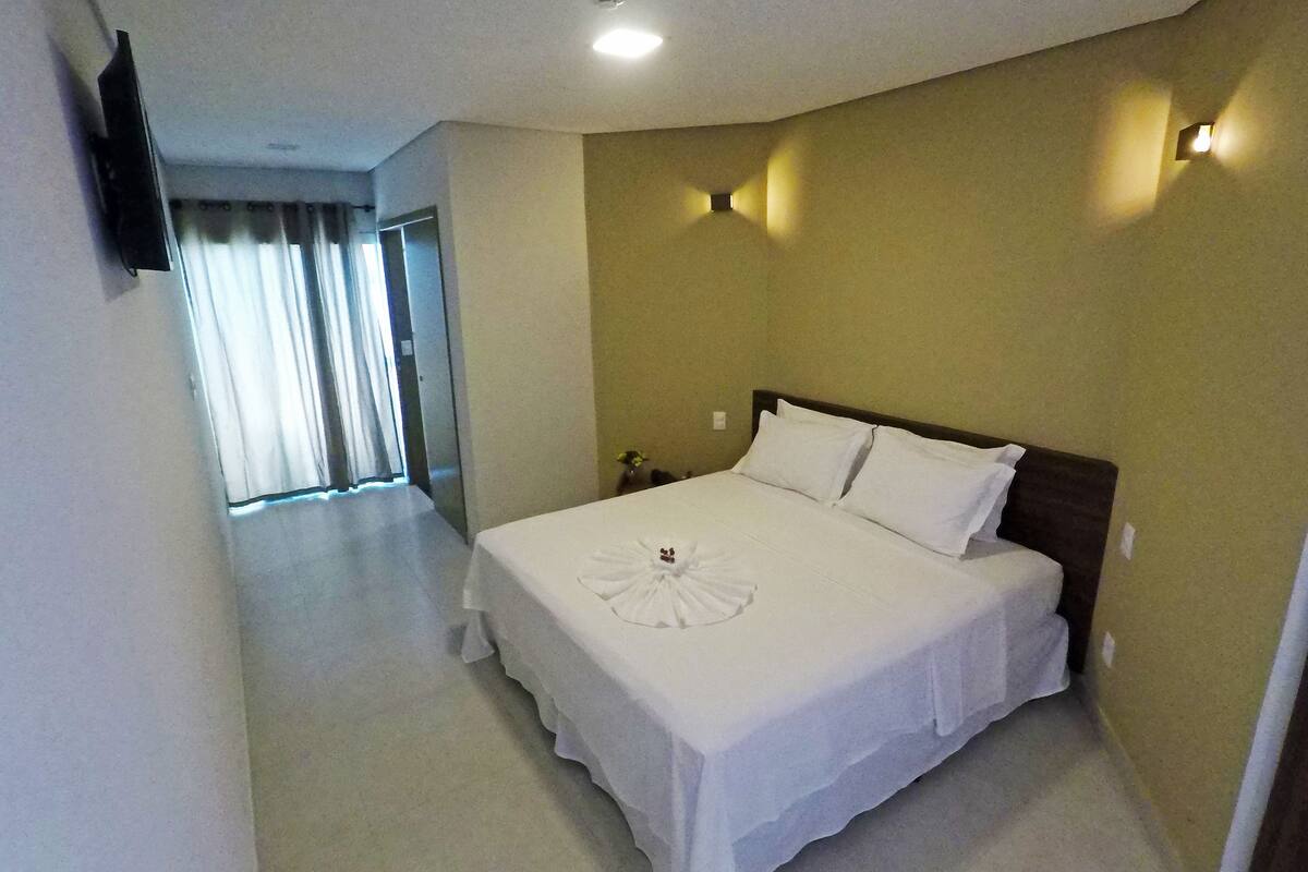 A hotel room with a large bed, a flat-screen TV mounted on the wall, and a window with curtains.