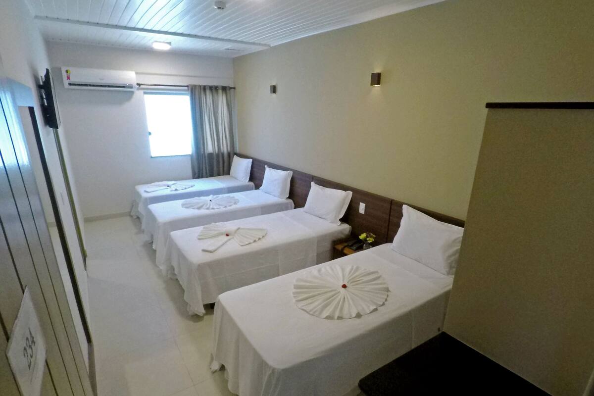 A hotel room with three single beds, each with a white bedspread and a decorative white towel. There is a small table with a vase of flowers, a nightstand, and a wall-mounted light.