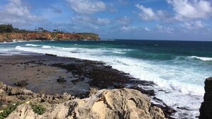 Une côte rocheuse où les vagues s’écrasent sur le rivage, un ciel dégagé et une étendue terrestre lointaine, couverte de végétation.