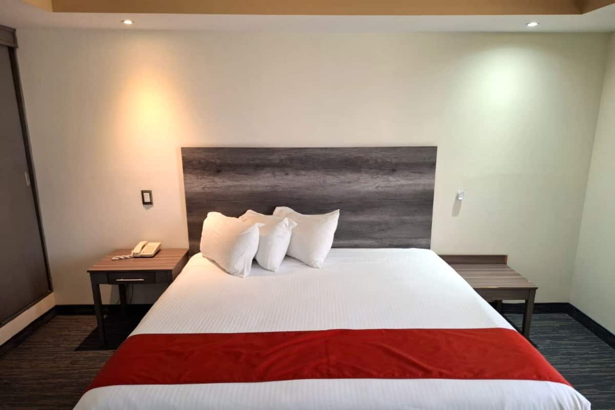 A hotel room with a large bed, a wooden headboard, a bedside table with a phone, and a red accent on the bedspread.