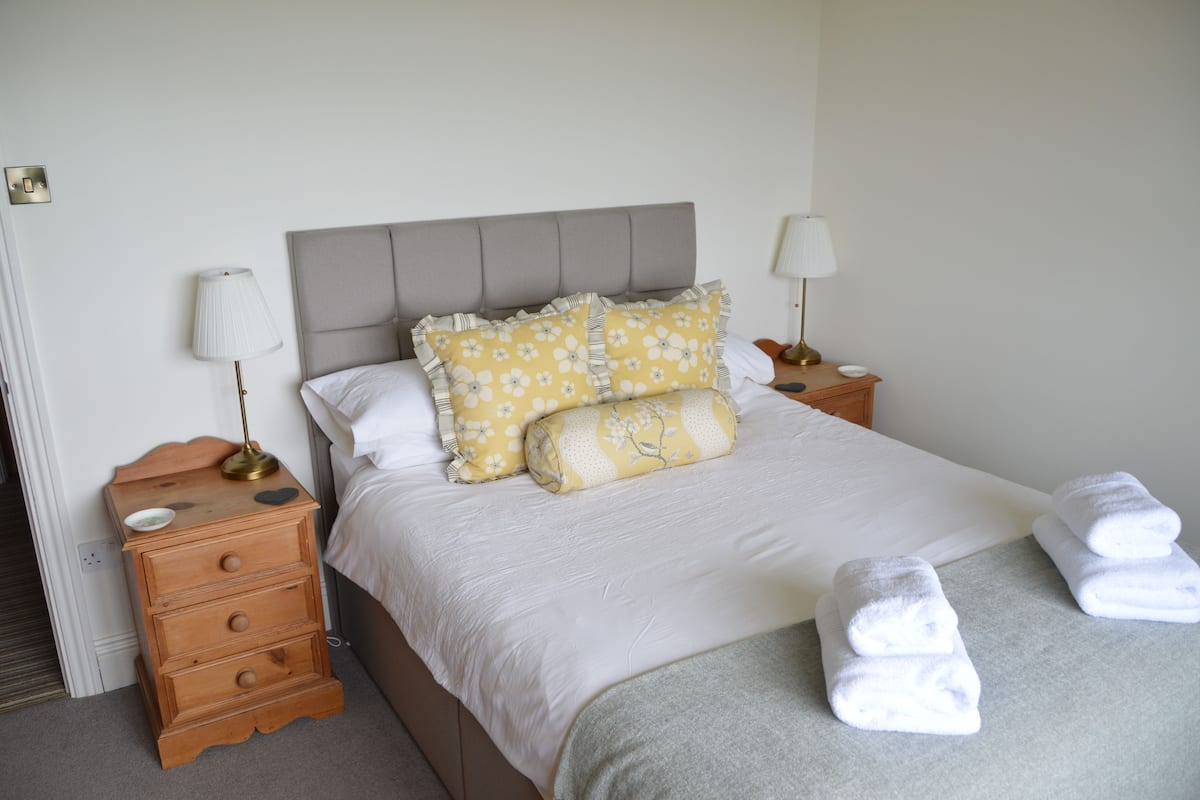 Un lit bien fait, avec une literie blanche, deux coussins décoratifs, une table de chevet avec une lampe et une petite commode.