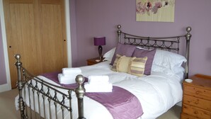A neatly made bed with white linens and purple accents, a wooden nightstand with a lamp, and a wooden dresser. - Trendrennen Farm B&B (Penzance)