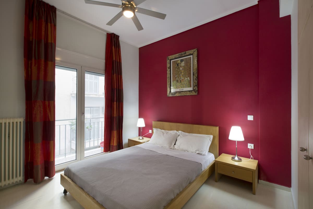 A bedroom with a bed, two bedside tables with lamps, a ceiling fan, a window with curtains, and a red wall with a framed picture.