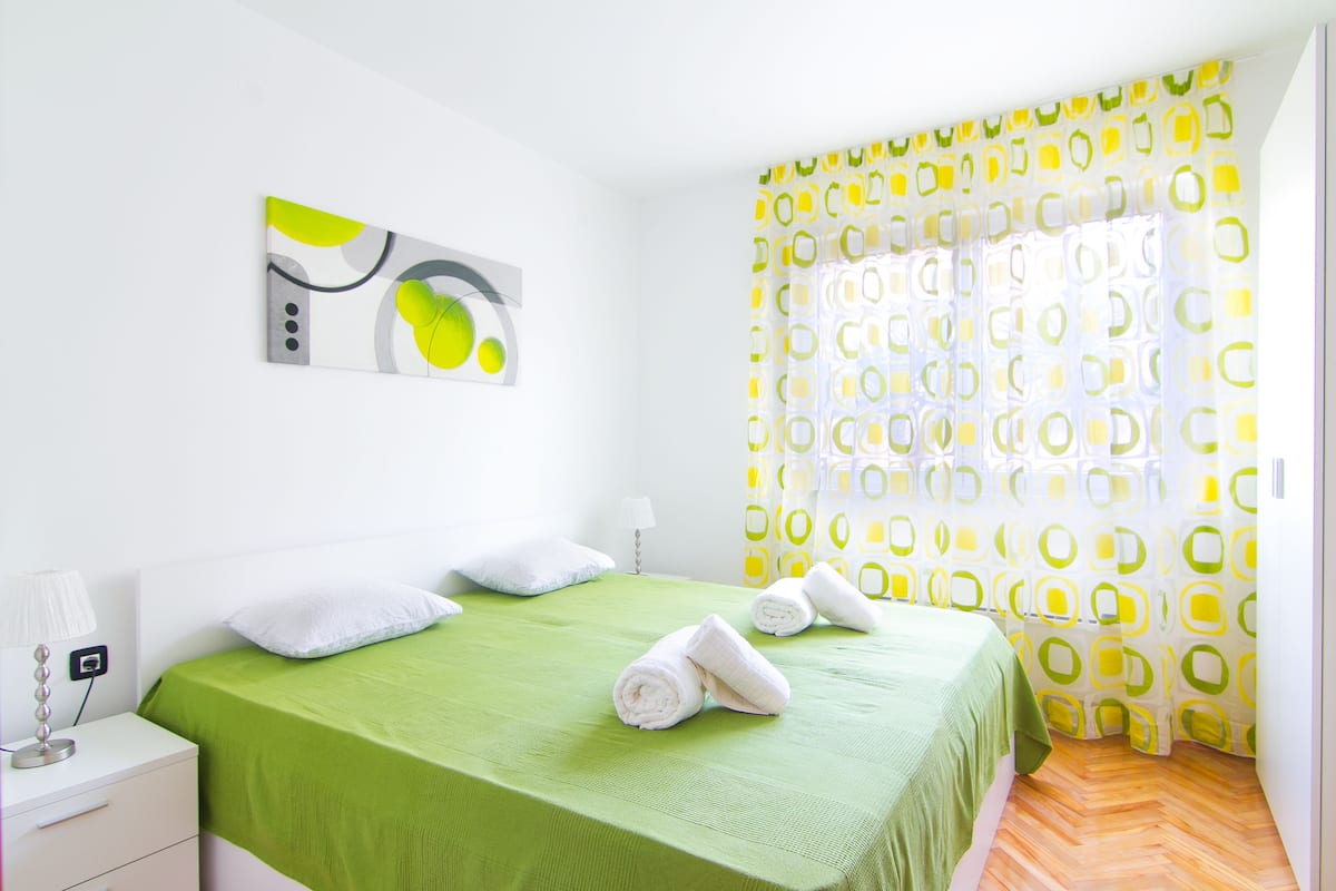 Una camera d'albergo con una coperta verde, cuscini bianchi, un quadro appeso al muro e una tenda decorata.
