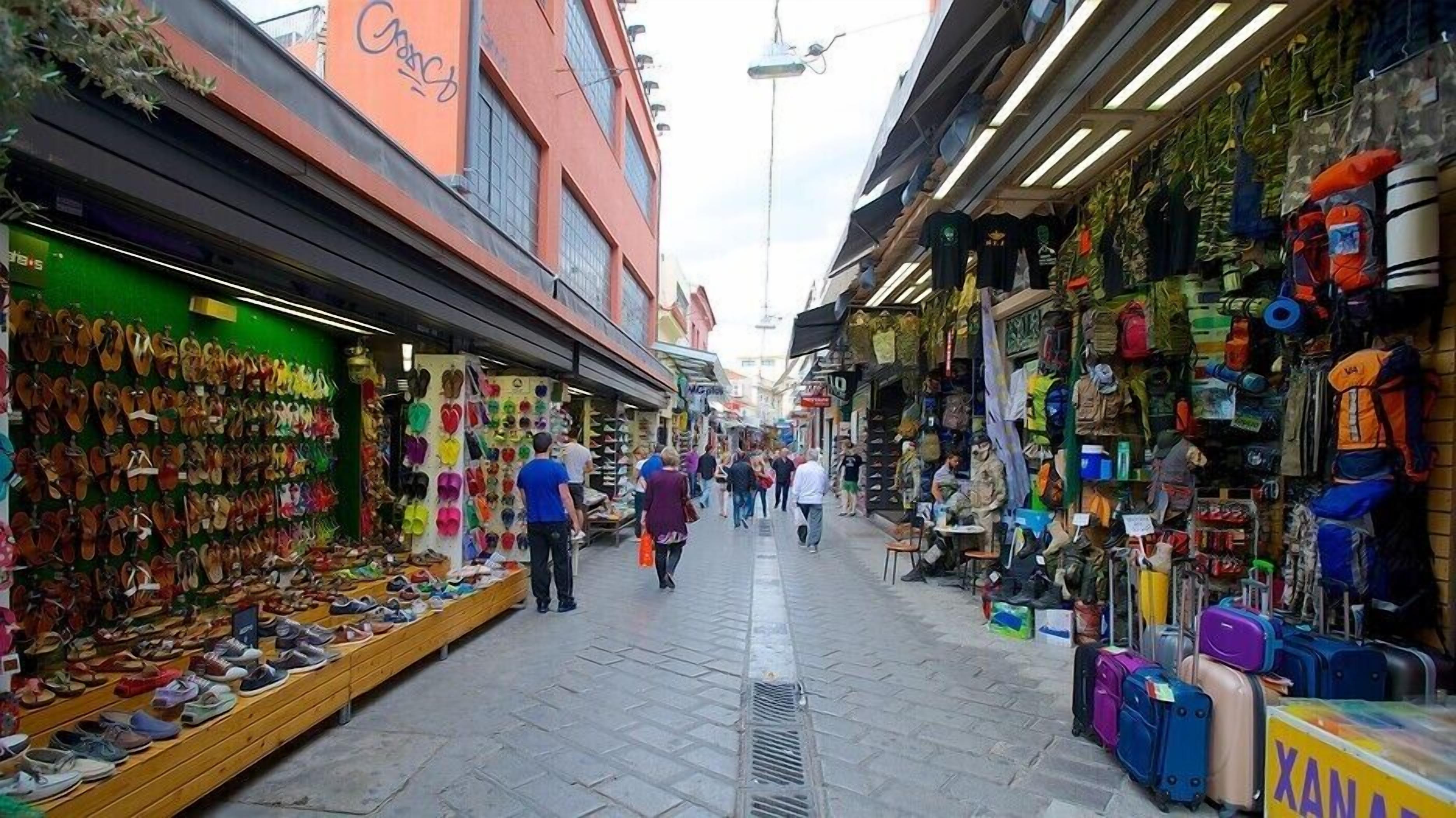 A narrow street with shops on both sides, including a shoe store and a store with luggage and camping gear.