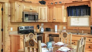 Interior de una cabaña de madera con una mesa de comedor para cuatro personas, una cocina con armarios y electrodomésticos de madera, y una araña.