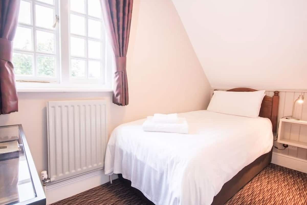 A bedroom with a bed, a radiator, a window with curtains, and a bedside table with a lamp.