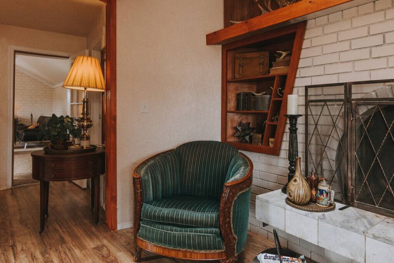 Um quarto com uma poltrona estofada em verde, uma mesinha de madeira, uma luminária e uma lareira com objetos decorativos.