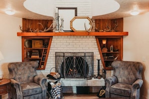 A cozy living room with a fireplace, two armchairs, and wooden shelves above the fireplace. - Sequim Lavender Castle (Sequim)