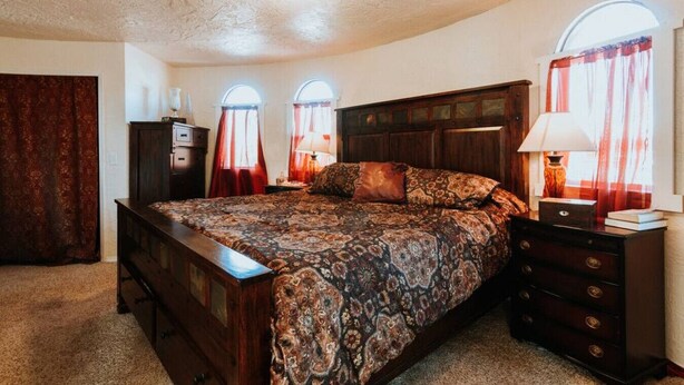 A bedroom with a large bed, two bedside tables, a dresser, and a wardrobe. There are three windows with curtains, and a ceiling light. - Sequim Lavender Castle (Sequim)