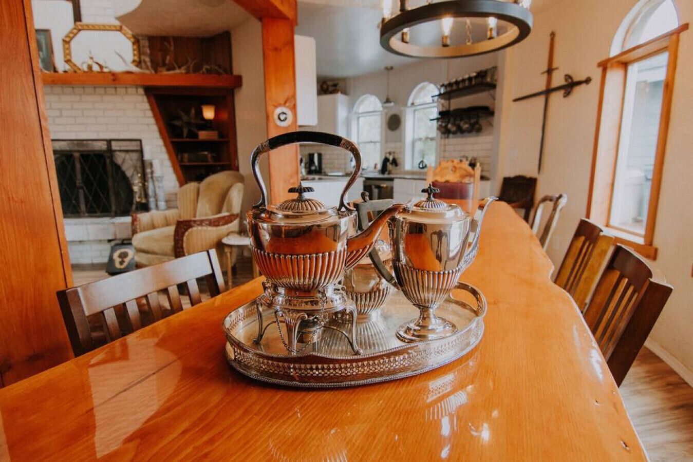 Een eettafel met een zilveren theeservies en een dienblad, in een kamer met een open haard, houten stoelen en een kroonluchter.