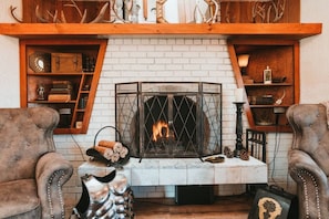 A fireplace with a white brick surround, flanked by wooden shelves with antlers and books. Two leather armchairs are positioned in front. - Sequim Lavender Castle (Sequim)