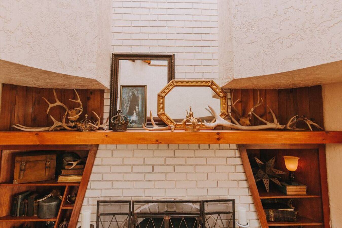 Una habitación con chimenea de ladrillo blanco, repisas de madera con ciervos y un espejo, además de un cuadro enmarcado sobre la chimenea.