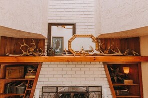 A room with a white brick fireplace, wooden shelves with antlers and a mirror, and a framed picture above the fireplace.