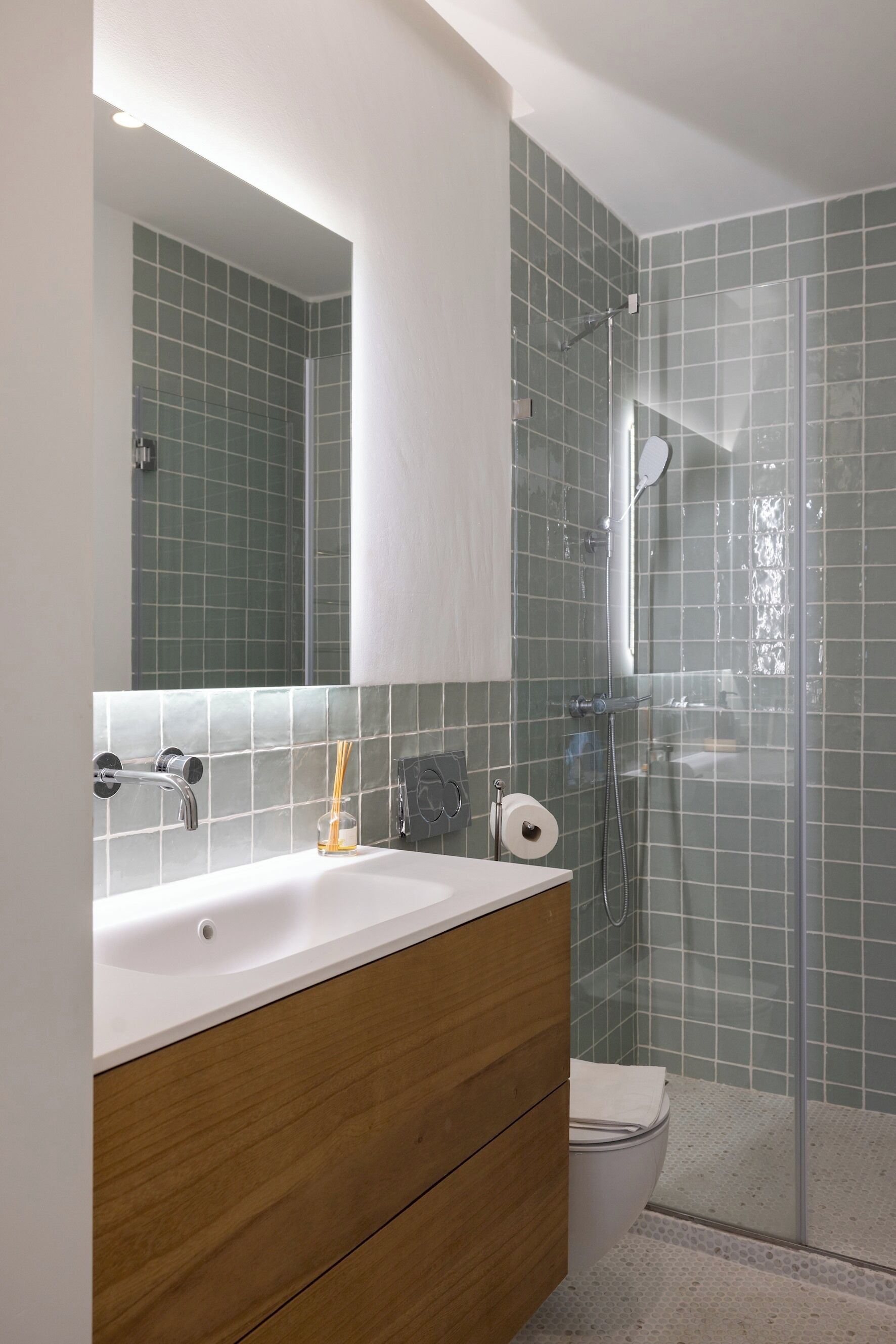 Une salle de bain avec un lavabo blanc, un meuble-lavabo en bois, une cabine de douche en verre et des murs recouverts de carreaux verts.