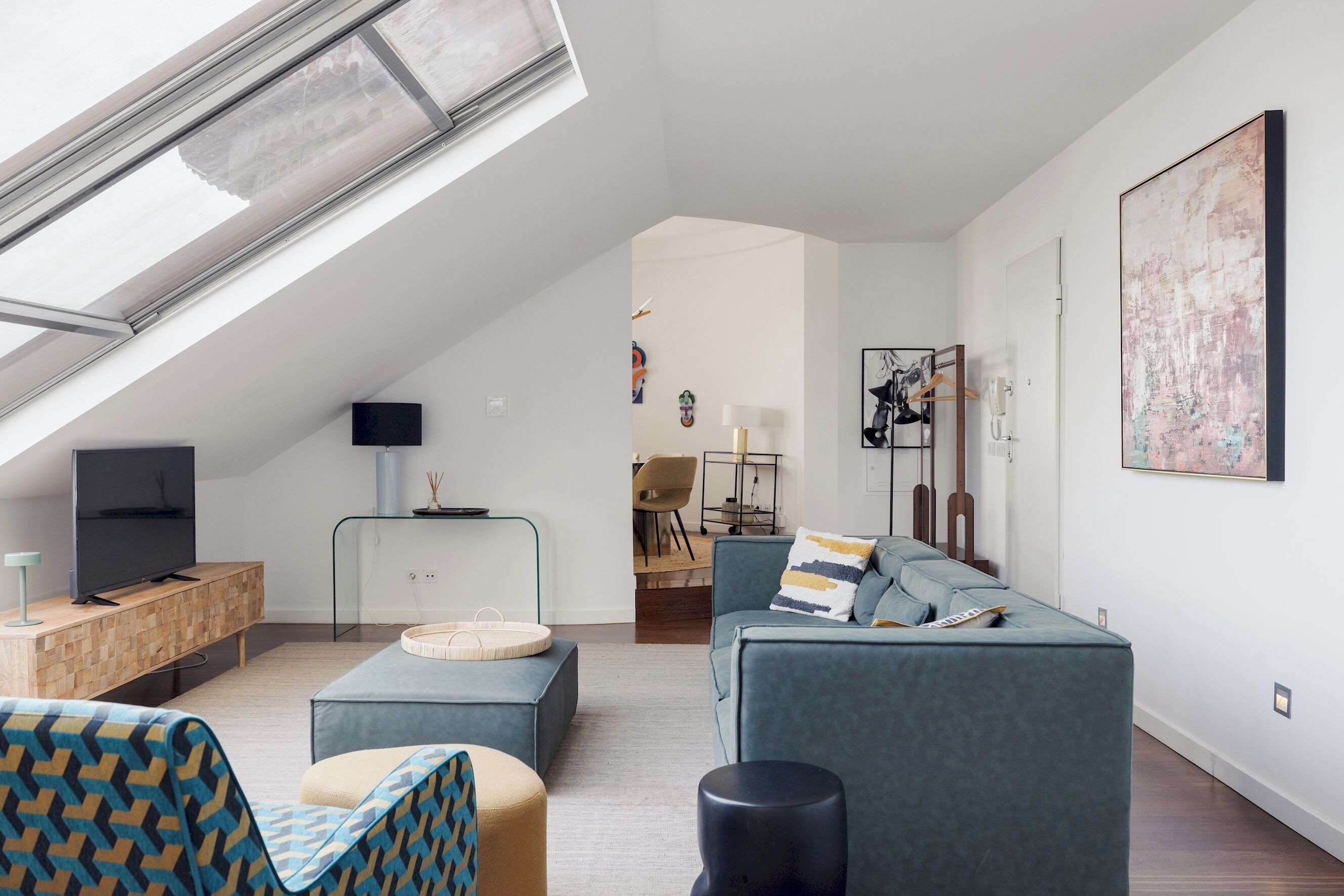 Ein modernes Wohnzimmer mit schrägen Wänden, einem Fernseher auf einer Holzkonsolle, einem Sofa, einem Sessel und einem Essbereich im Hintergrund.