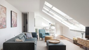 Un appartement moderne de style « loft », avec un plafond haut, une lucarne, un canapé gris, un fauteuil à motifs bleus et un téléviseur posé sur une console en bois.