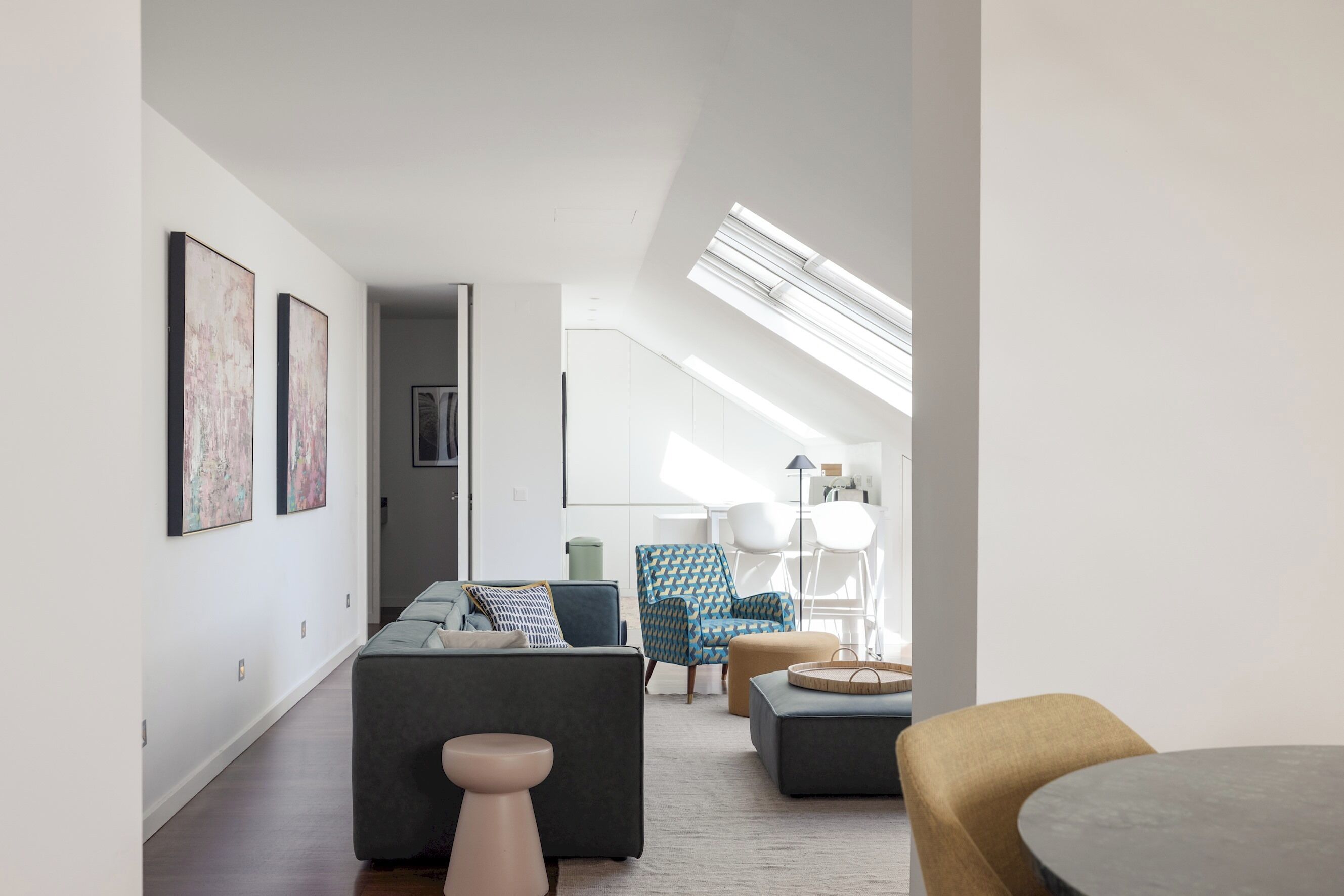 A modern living room with a sofa, ottoman, and armchair, a round table, and a lamp. There are framed artworks on the wall and a skylight above.