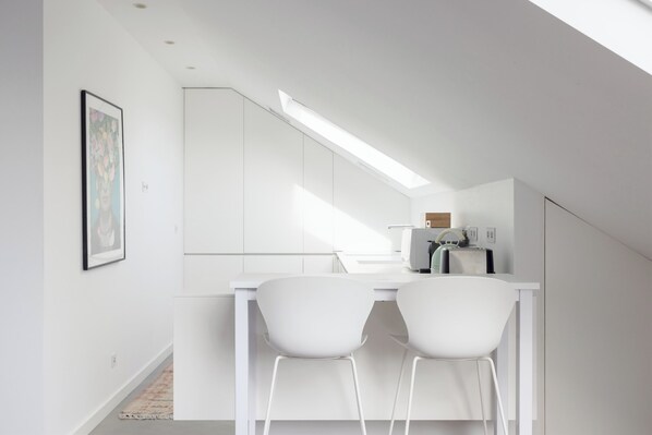 Cocina moderna con armarios blancos, un tragaluz y dos sillas blancas junto a una mesa blanca.
