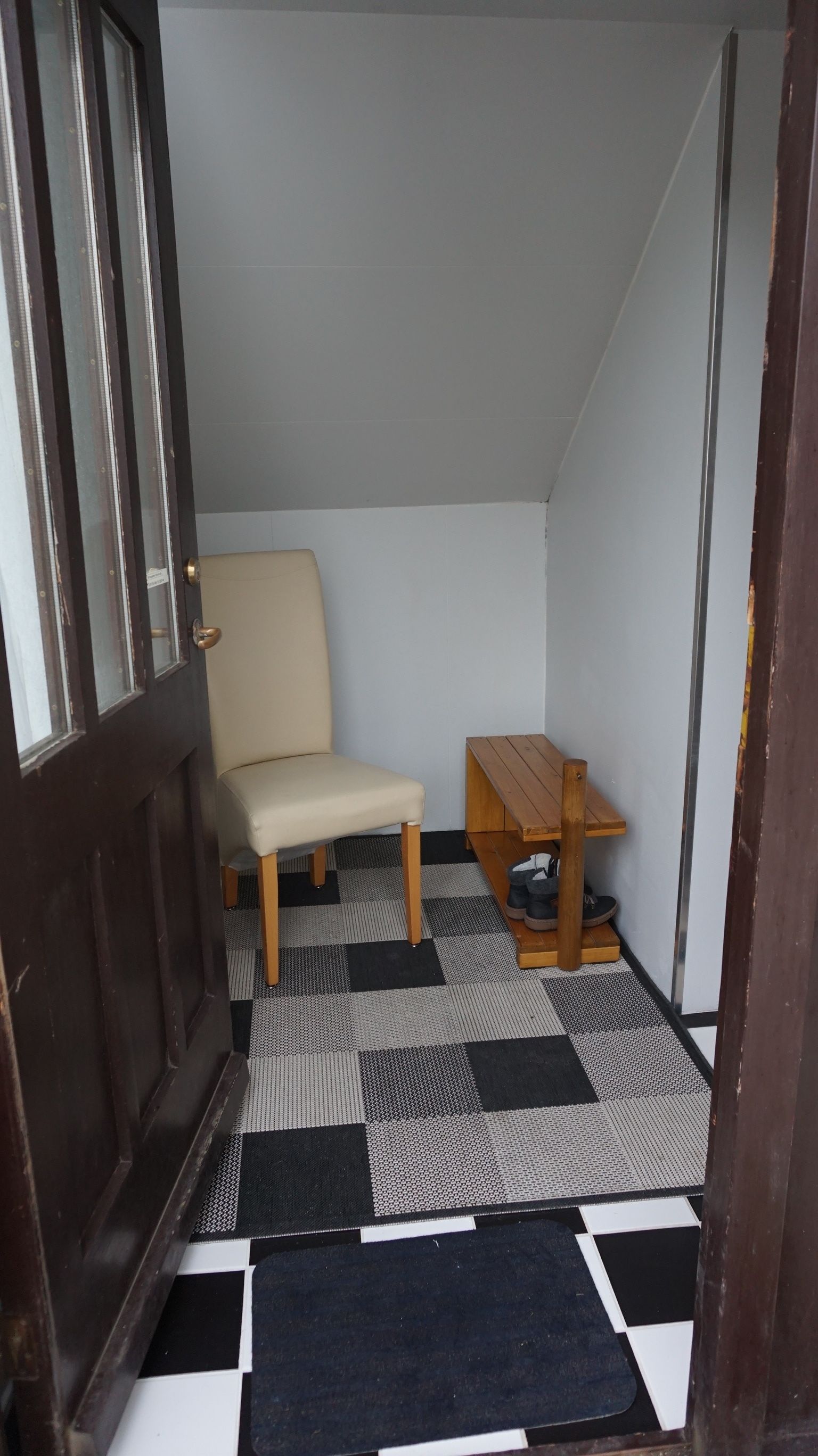 Une petite pièce fermée, dotée d’un sol à carreaux, d’une table en bois, d’une chaise et d’une porte avec une poignée en métal.