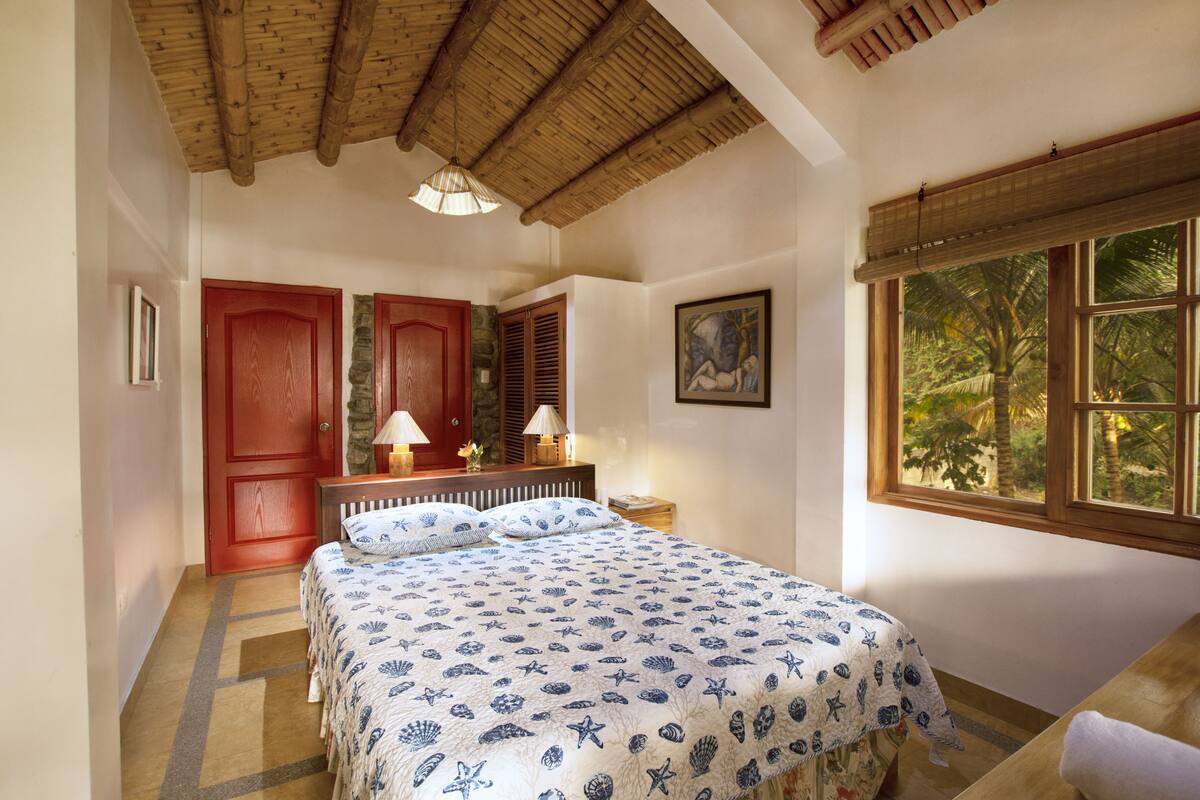A bedroom with a bed, a wooden ceiling, a window with a view, and a door.