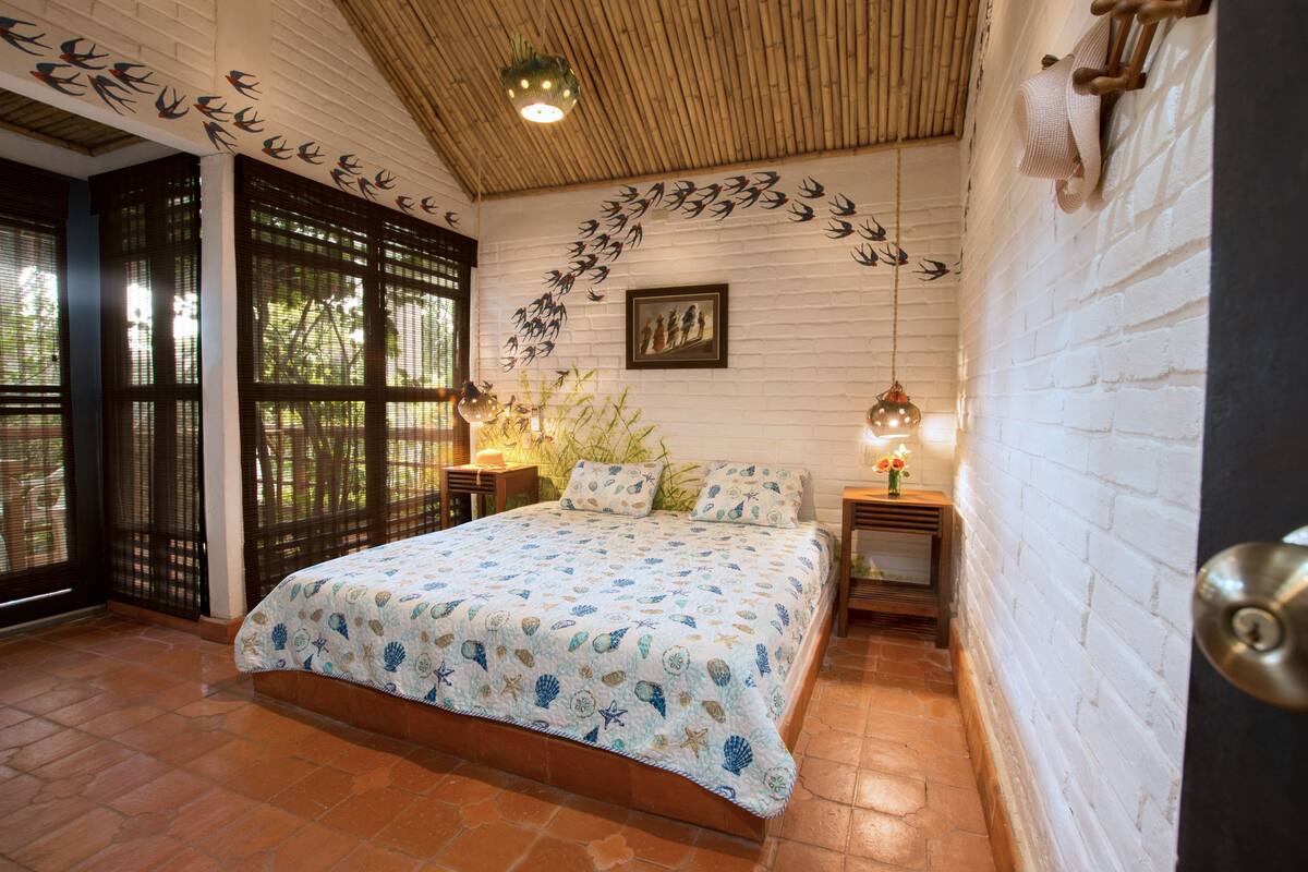 A bedroom with a bed, a wooden ceiling, a white brick wall, and a window with a view of trees.