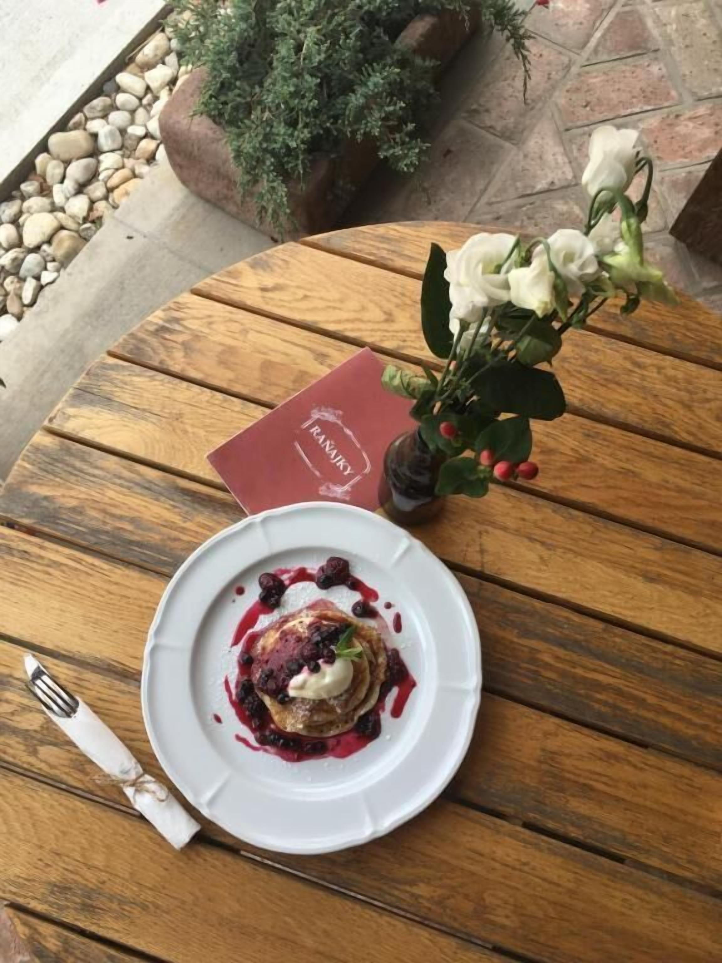 A wooden table with a plate of pancakes topped with berries and cream, a vase with white flowers, and a red menu.
