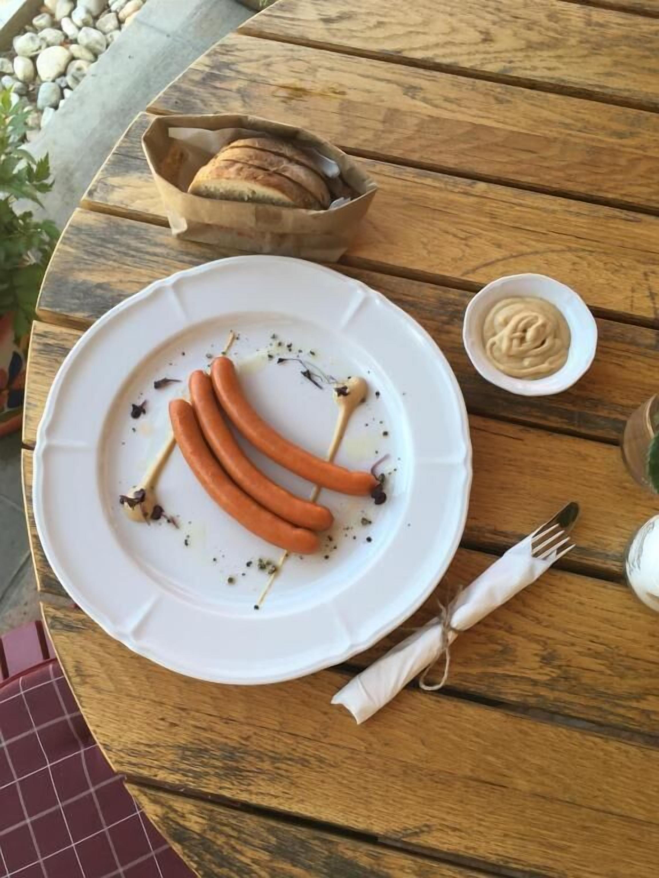 Une table en bois sur laquelle se trouve une assiette blanche remplie de saucisses, un petit bol de moutarde et un panier de pain.