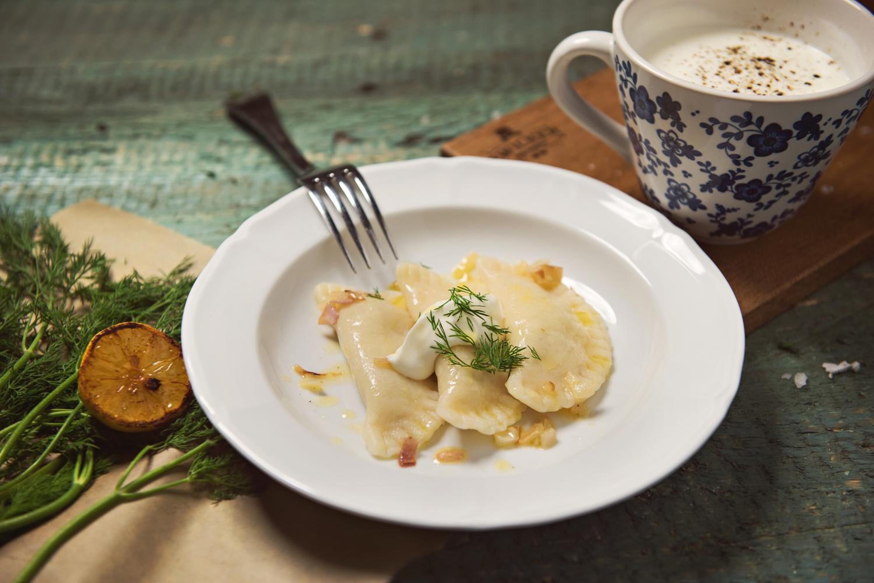 A plate of pierogi with a fork, a cup of coffee, and some herbs on a rustic table.