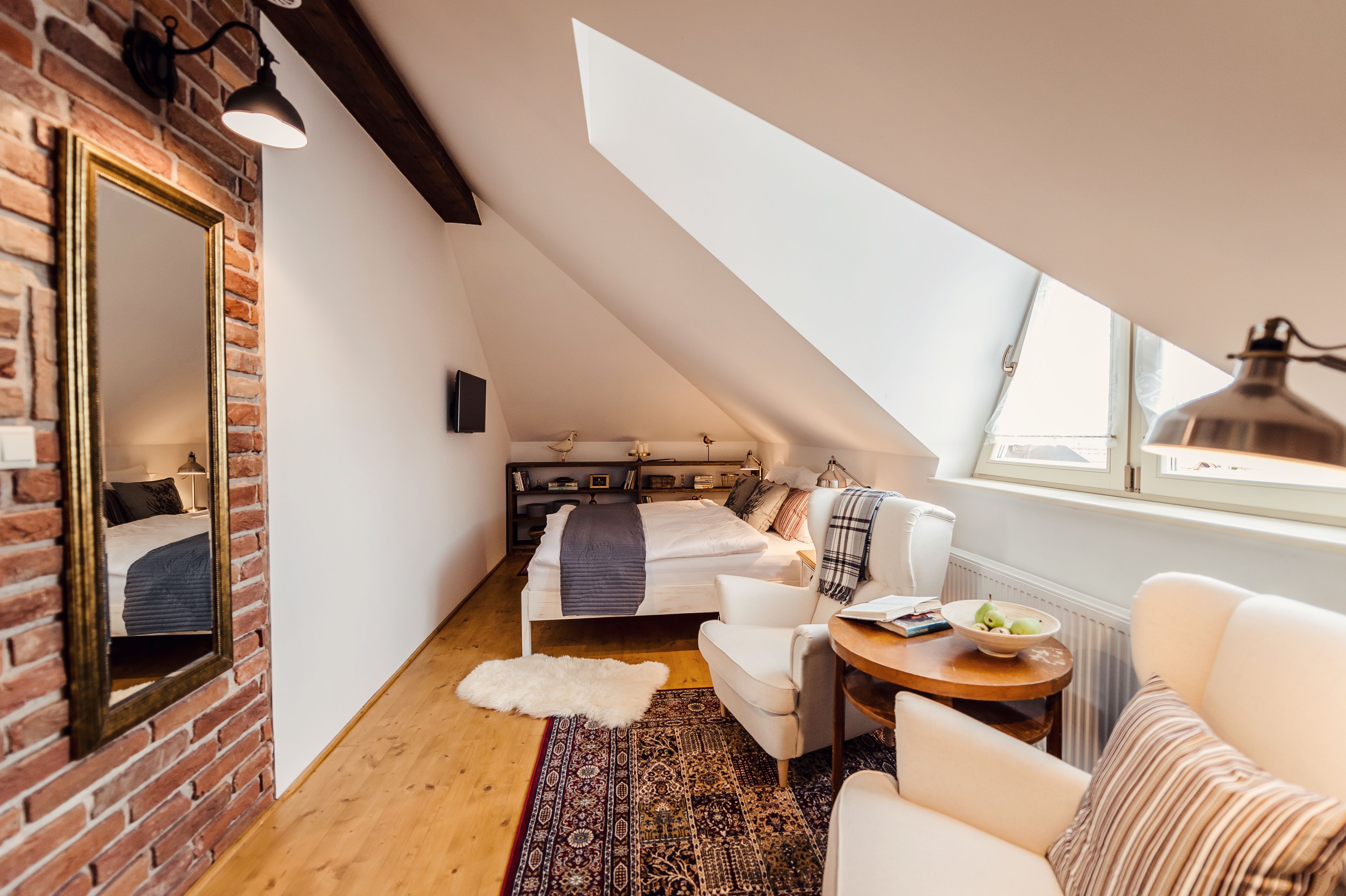 A cozy loft bedroom with a sloped ceiling, exposed brick walls, a large mirror, a bed with a white headboard, a small white sofa, a round wooden table, and a view of the sky through a skylight.