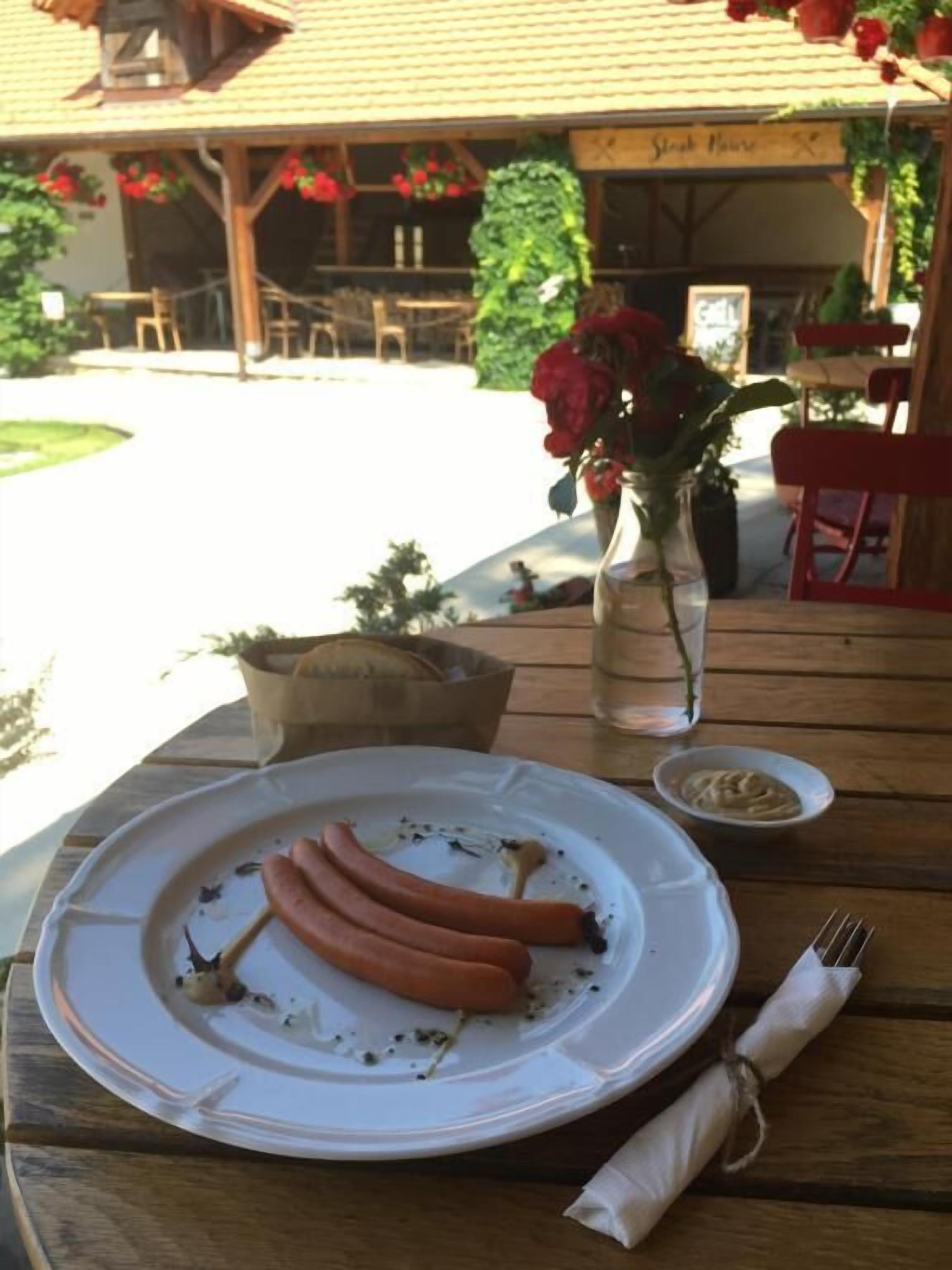 A dining table with a plate of sausages, a small bowl of mustard, and a vase with red flowers.