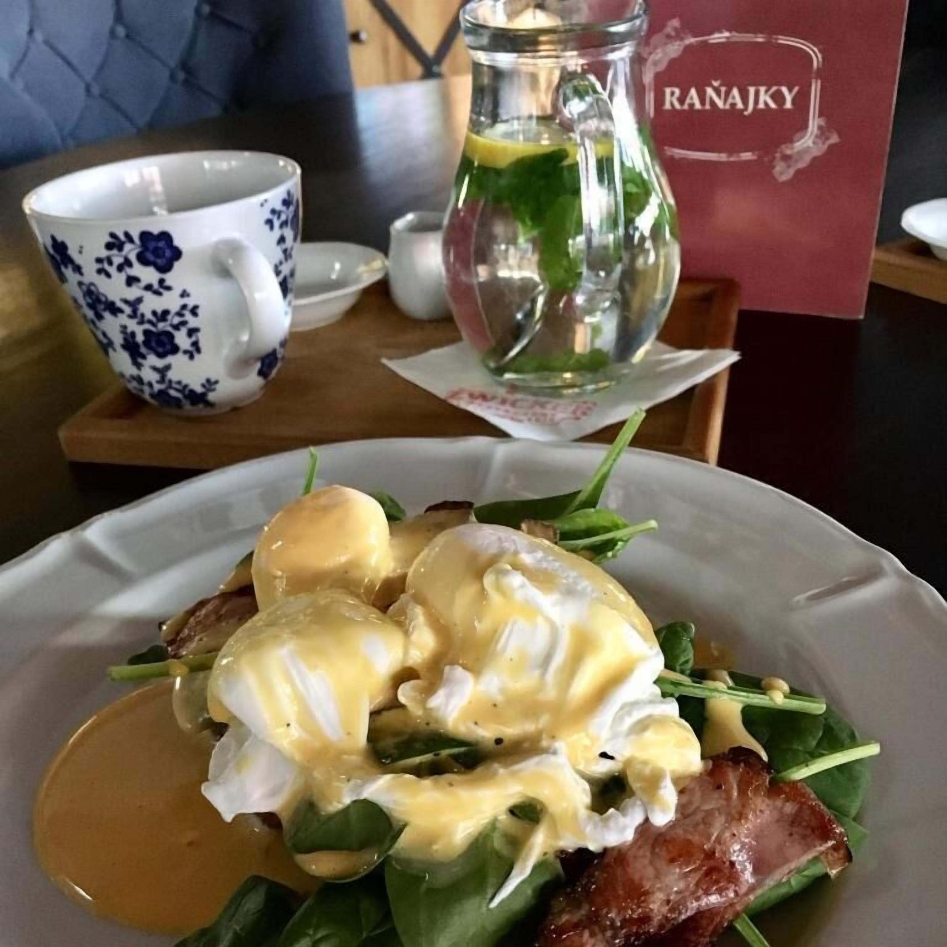 Un piatto con uova alla Benedict, pancetta e spinaci su un tavolo, con una tazza azzurra e bianca e un cartello rosso.
