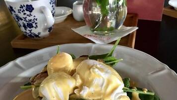 Un piatto con uova alla Benedict, pancetta e spinaci su un tavolo, con una tazza azzurra e bianca e un cartello rosso.