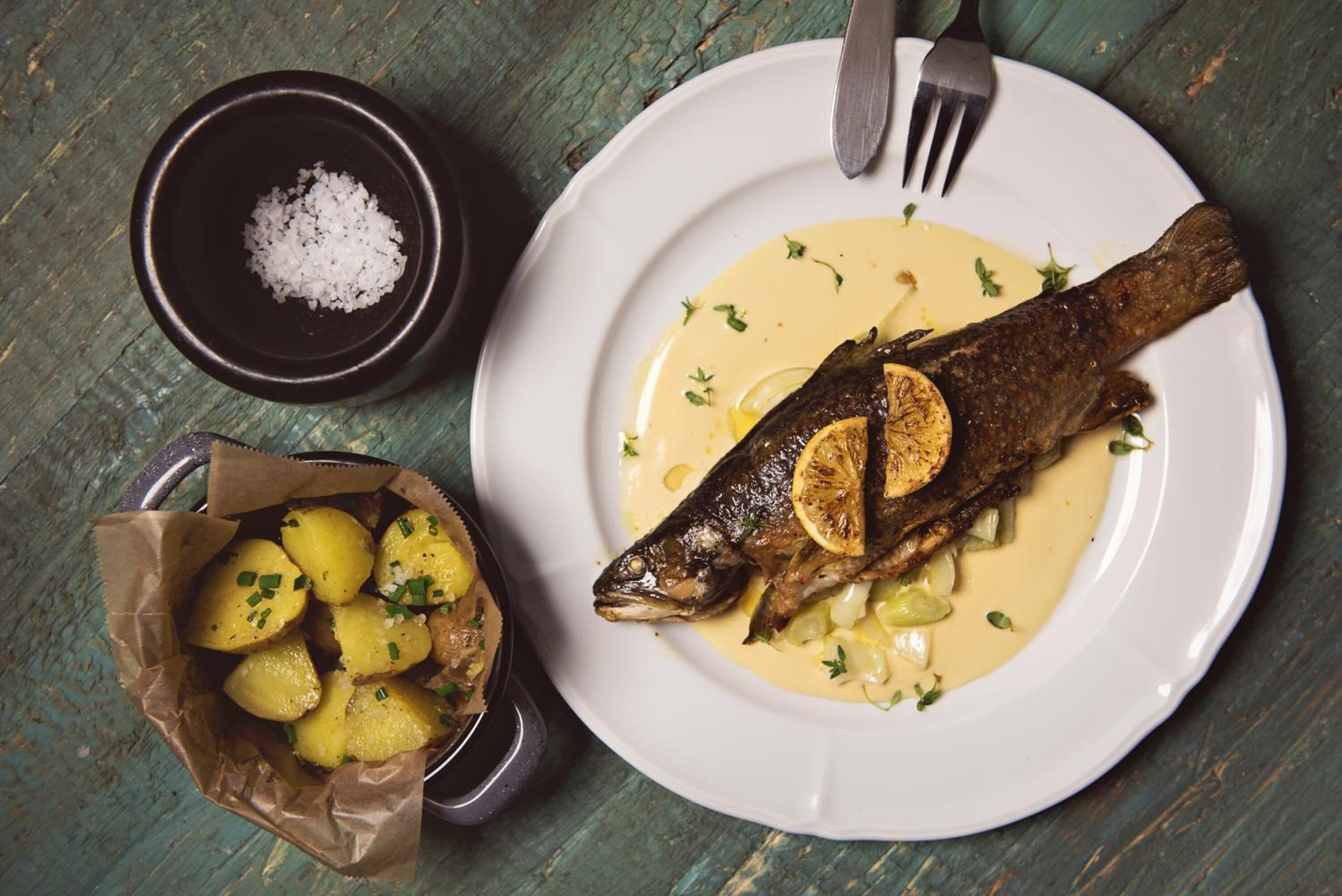 A plate with a fish, lemon slices, and a creamy sauce, accompanied by a bowl of potatoes and a small bowl of salt.