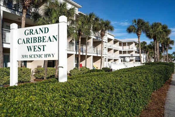 Ett hotellskylt för Grand Caribbean West, beläget på 3191 Scenic Hwy, som visar en byggnad med flera våningar, balkonger och palmer.