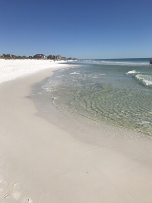 En strand med klart vatten, vit sand och några få byggnader i bakgrunden.