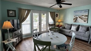 A living room with a dining area, a ceiling fan, a television, and a view of the outdoors through large windows. - Fantastic Beach location! With Ocean View.1 Bedroom, Bunk beds, Sofa bed 1 bath. (Destin)