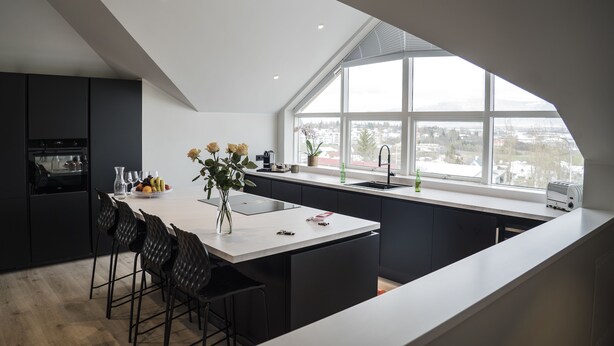 A modern kitchen with a large island, dark cabinetry, and a view of a cityscape through a large window. - Óm Apartments (Reykjavik)
